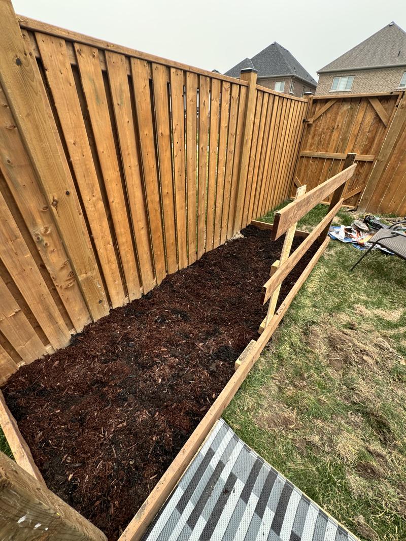A newly built garden bed edged with timber running along the base of a wooden fence, soil levelled and ready for planting.